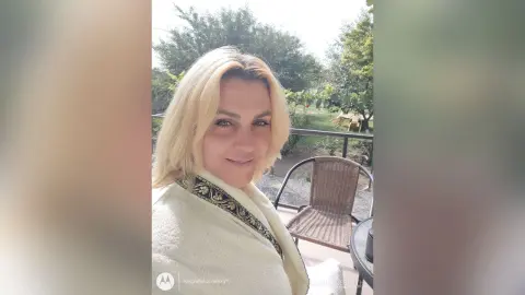Smiling blonde woman in a white textured sweater relaxing on a sunny outdoor patio.