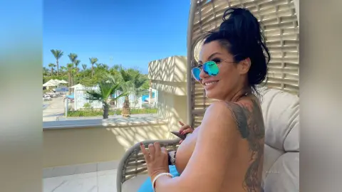 MissyJolie smiles while sitting outdoors in a chair, wearing sunglasses and showing her bare back and shoulder with a large dark tattoo.