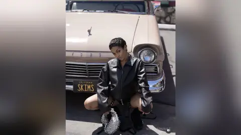 Stylish woman in a black leather jacket sitting by a vintage beige Ford car in bright sunlight.