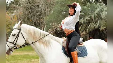 FlorenciaCopper sits upright on a white horse wearing a white blouse and black leggings, wearing a brown cowboy hat.