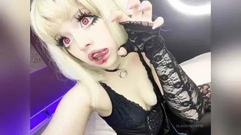 Gothic girl with blonde hair, red eyes, and black lace outfit posing in a moody bedroom setting.