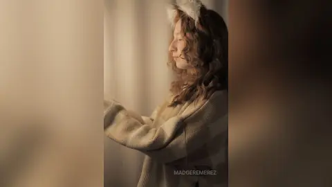 Dreamy portrait of a young woman in a cozy cream sweater standing by a window in soft, warm sunlight.