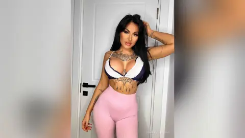 Tattooed woman in a white and purple bikini top and pink leggings posing indoors with a confident mood.