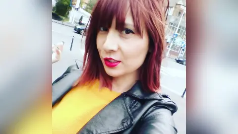 AryaSin wearing a black leather jacket and yellow top with red lipstick and red hair, posing outdoors on a street.