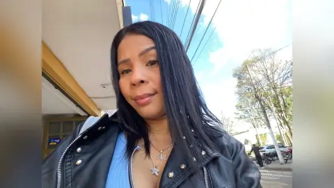 Smiling woman in a black leather jacket and blue top outdoors under a bright blue sky.