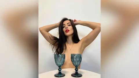 Brunette woman with red lips posing behind blue glass goblets in a minimalist white setting.