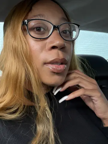 LexBright wearing black glasses and a black top, posing with her hand near her face and long white nails.