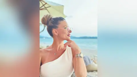 Smiling woman in a white one-shoulder top and sunglasses relaxing on a sunny tropical beach.
