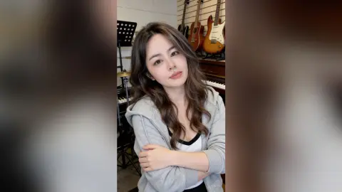 Young woman in a grey hoodie smiling in a music studio surrounded by guitars.