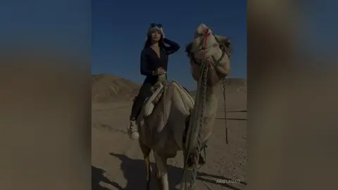 Stylish woman in black outfit riding a camel through a sunny desert landscape.