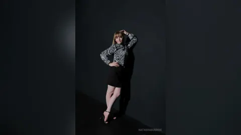 NatashaSunrise — NatashaSunrise poses in a black and white striped long-sleeve shirt and black skirt against a dark background.