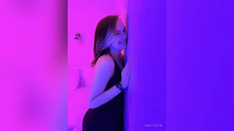 EmilyGiffin smiles while leaning against a wall in a black tank top, lit by bright pink and purple neon lights.