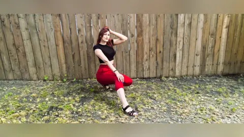 Redheaded woman in a black t-shirt and red pants posing against a rustic wooden fence in a sunny garden.