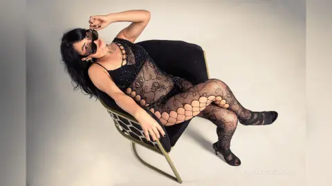 Glamorous woman in a black lace bodysuit and tights relaxing in a velvet chair against a white studio background.