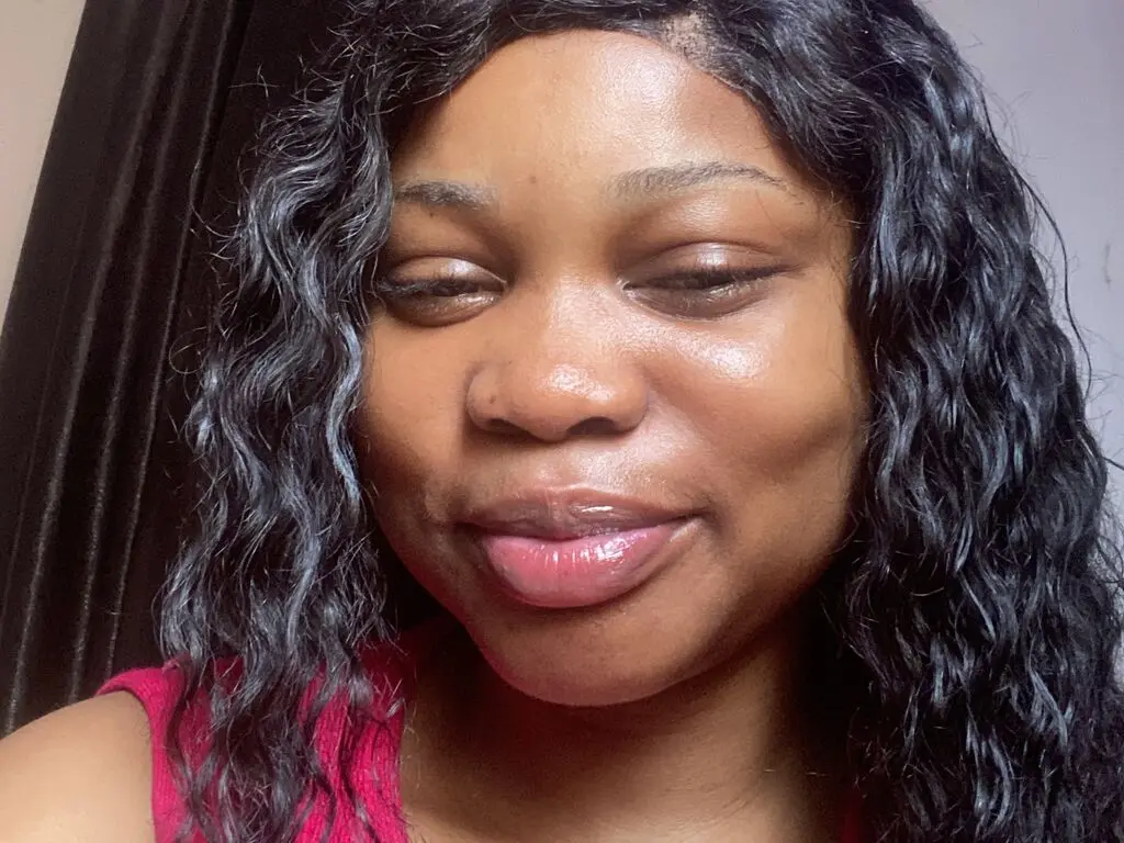 Smiling Black woman with long wavy black hair wearing a pink top in a cozy indoor setting.