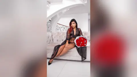Elegant woman in a black sheer lace robe sitting on a white sofa holding a bouquet of red roses.