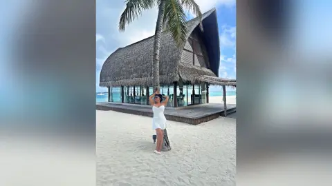 MissyJolie stands on a white sand beach wearing a white mini dress, posing behind a palm tree near a thatched hut.