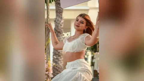 Smiling young woman in a white lace crop top and skirt sitting on a swing outdoors in bright sunlight.