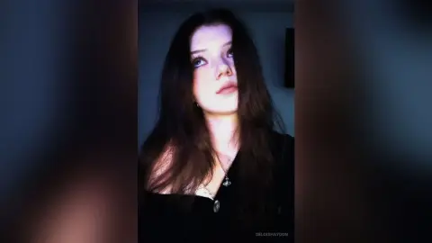 Young woman with long dark hair wearing a black top and silver necklace in moody, dim lighting.