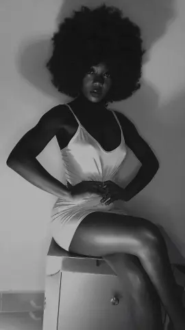 Moody black and white portrait of a woman with an afro wearing a white slip dress sitting on a cabinet.