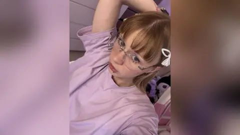 Young woman with blonde bangs and glasses wearing a lilac t-shirt in a cozy indoor setting.