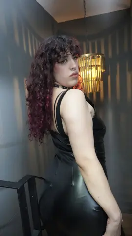 Woman with curly red hair in a black sleeveless dress posing in moody, dramatic indoor lighting.