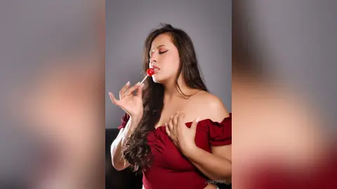 Beautiful woman in a red off-the-shoulder top eating a red lollipop against a grey studio background.