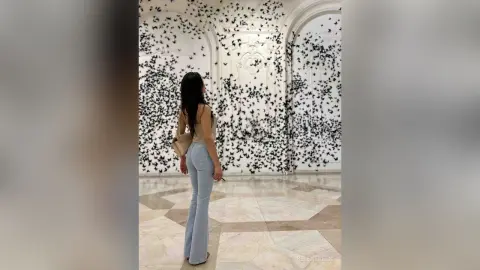 Woman in light blue flared jeans and tan top standing before a dreamy black butterfly wall installation.