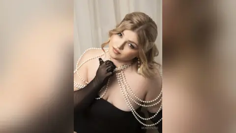 Elegant woman in a black dress and pearl body jewelry posing glamorously against a white backdrop.
