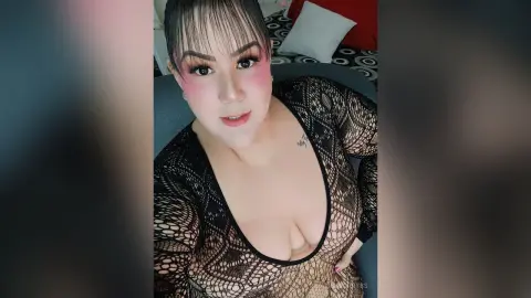 Curvy woman with pink makeup wearing a black lace bodysuit in a cozy bedroom setting.