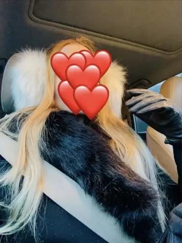 Blonde woman in a black winter coat with a fluffy faux fur collar sitting in a car.