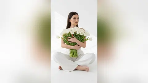 ZenyaSin sits cross-legged against a white background, holding a large bouquet of white tulips against her chest with her long dark hair down.