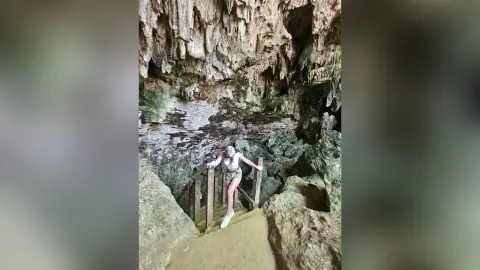 RossiNoleen stands on a wooden staircase inside a rocky cave, wearing a white bikini top and dark bottoms.