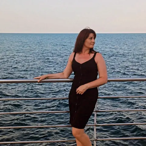 MidnightSecretX stands by a metal railing overlooking the ocean, wearing a black sleeveless button-down dress with her brown hair down.