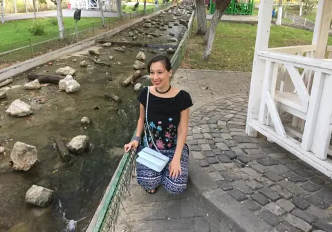 Smiling woman in a black floral top and patterned pants sitting by a scenic stone stream in a park.