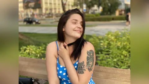 Smiling young woman with long dark hair and owl tattoo sitting on a park bench in soft sunlight.