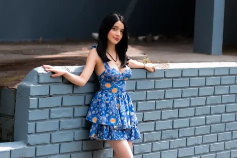 Young woman in a blue floral sundress leaning against a grey brick wall in bright daylight.