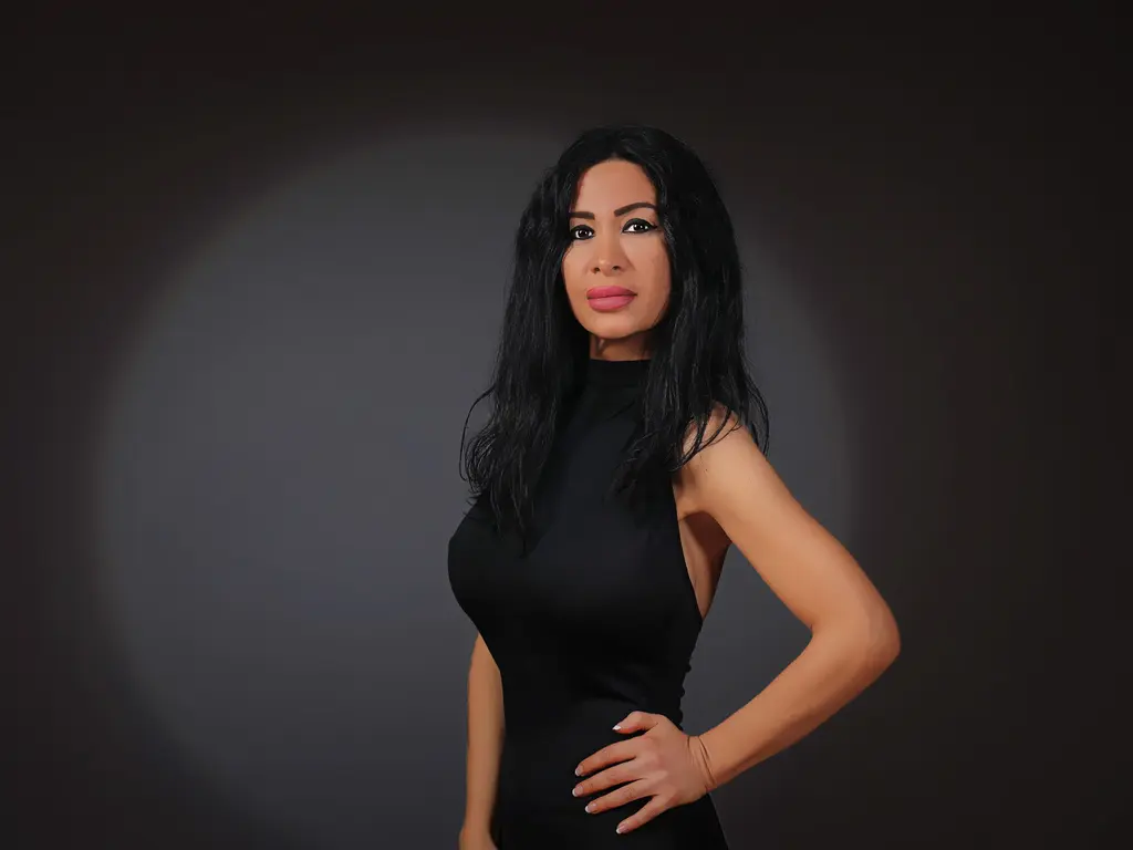 Elegant woman in a sleek black sleeveless dress posing against a dark studio background with moody lighting.