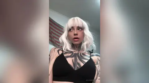 Young woman with white hair and tattoos wearing a black crop top in a moody indoor setting.