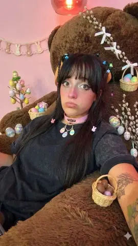 Young woman with dark hair and pink choker sitting in front of a giant teddy bear decorated with Easter eggs.