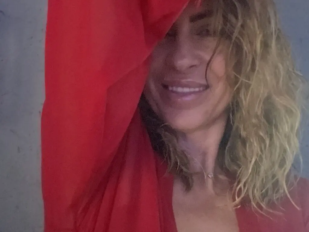 Smiling woman with wavy blonde hair draped in a vibrant red fabric against a neutral background.