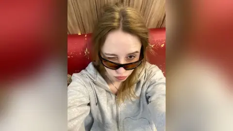 Blonde woman in a grey hoodie and sunglasses posing in a red booth for a casual selfie.