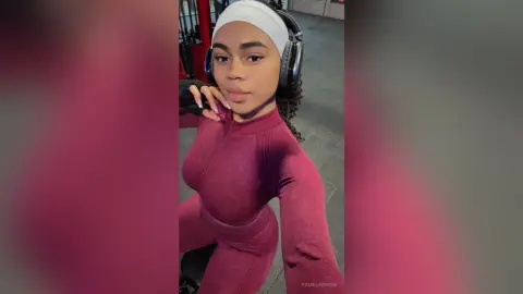 PamelaCreed wears a white headband and black headphones while posing in a tight, maroon long-sleeve top and matching leggings.