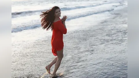 Happy woman in a red summer dress walking through ocean waves on a sunny beach.