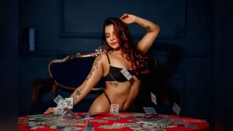 SofiaGreco sits at a red table covered in playing cards, wearing black lingerie and showing off her tattoos.