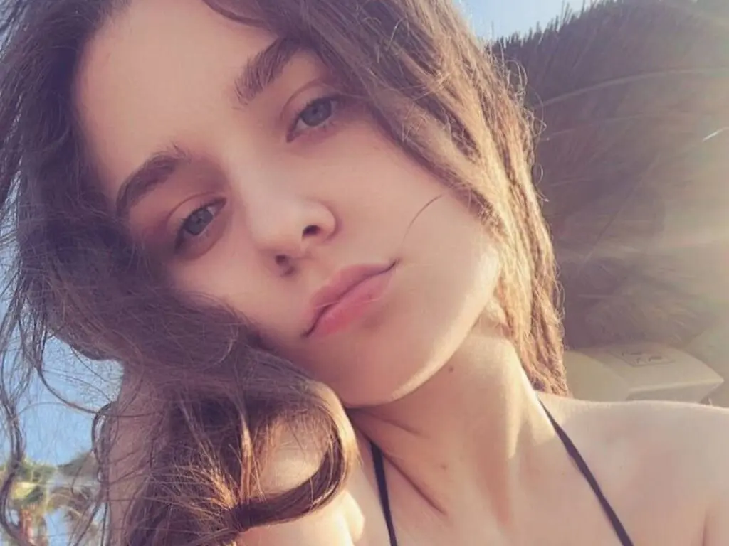 LuanaContratto poses close-up with long wavy brown hair and a black string strap visible against a bright outdoor background.
