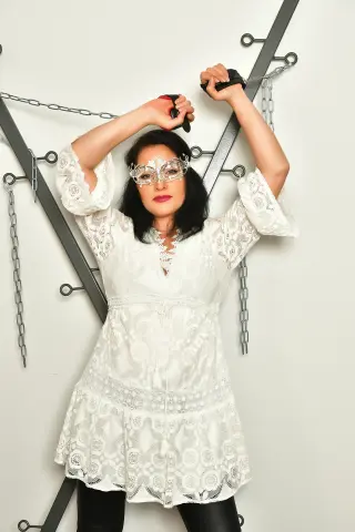 Woman in white lace dress and masquerade mask posing against a minimalist gray background.