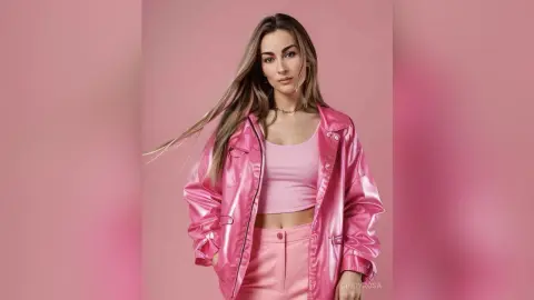 Stylish woman in a pink leather jacket and crop top posing against a soft pink background with a confident mood.