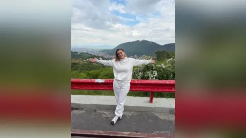 AnishaBee stands outdoors against a mountain landscape, wearing a white long-sleeved sweatshirt and matching white pants with her arms spread wide.