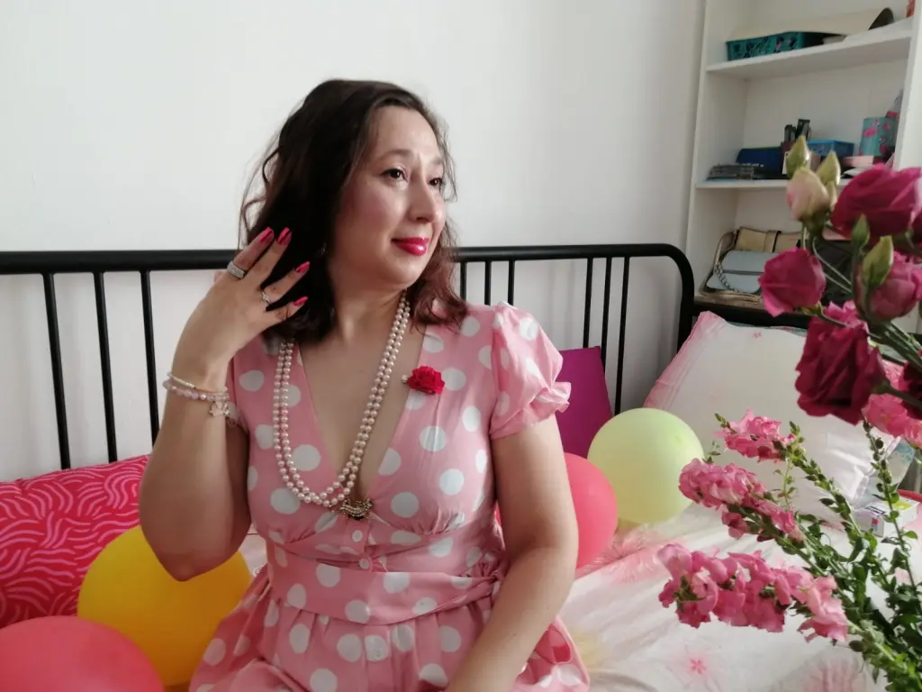 LadyDaphnne smiles while wearing a pink polka-dot top and a white pearl necklace, holding her hand near her face with red manicured nails.